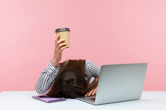 Woman feeling tired at work with head resting on the desk while one hand holding a paper cup coffee