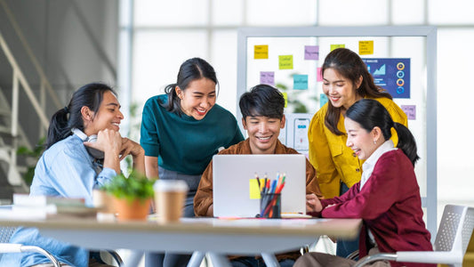 5 young asian man and woman huddling together together over a laptop in a startup office, happy in discussion.