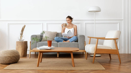 A young, caucasian woman relaxing on a couch in a minimalist living room. She has a book on one hand and a cup of beverage on the other. She appears to be in contentment as she relaxes in the peaceful and serene surrounding. 