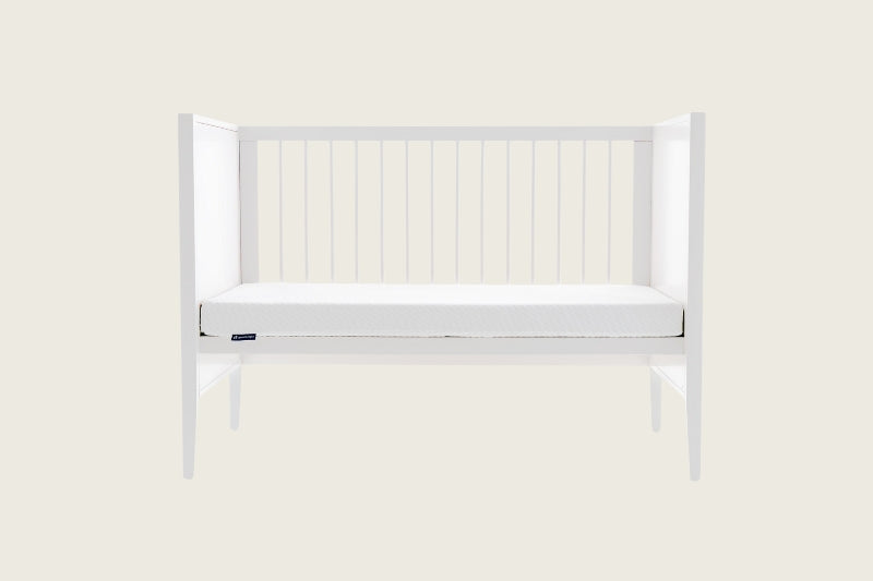 The Good Knight Aelia baby cot mattress placed in a white baby cot. The front rail of the baby crib is removed, showing the Aelia mattress laying flat on its bed base. 