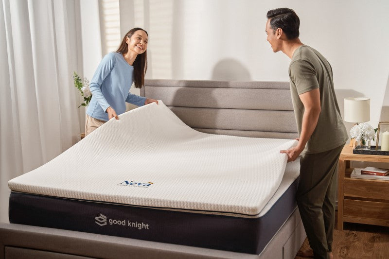 A couple setting up their Neva mattress topper from Good Knight onto their Good Knight mattress. its comfortably on a Good Knight mattress. The woman gazes lovingly at her partner as he smiles back, both enjoying a moment of relaxation and connection.