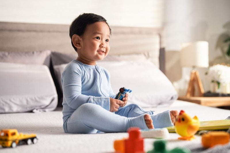 A smiling toddler sits on a Good Knight mattress, surrounded by toys