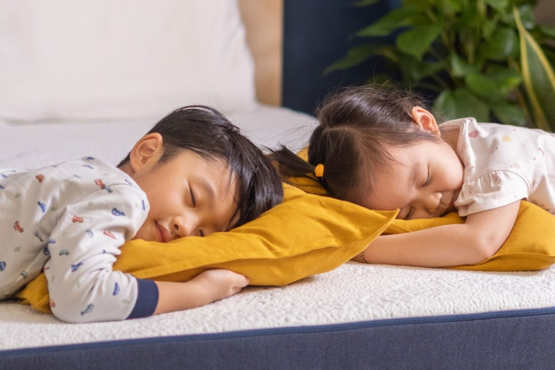 A little boy and a girl are sleeping on a Good Knight Mattress with a pillow and some green plants in the background. The children are sleeping peacefully and the boy even have a little smile while sleeping. 